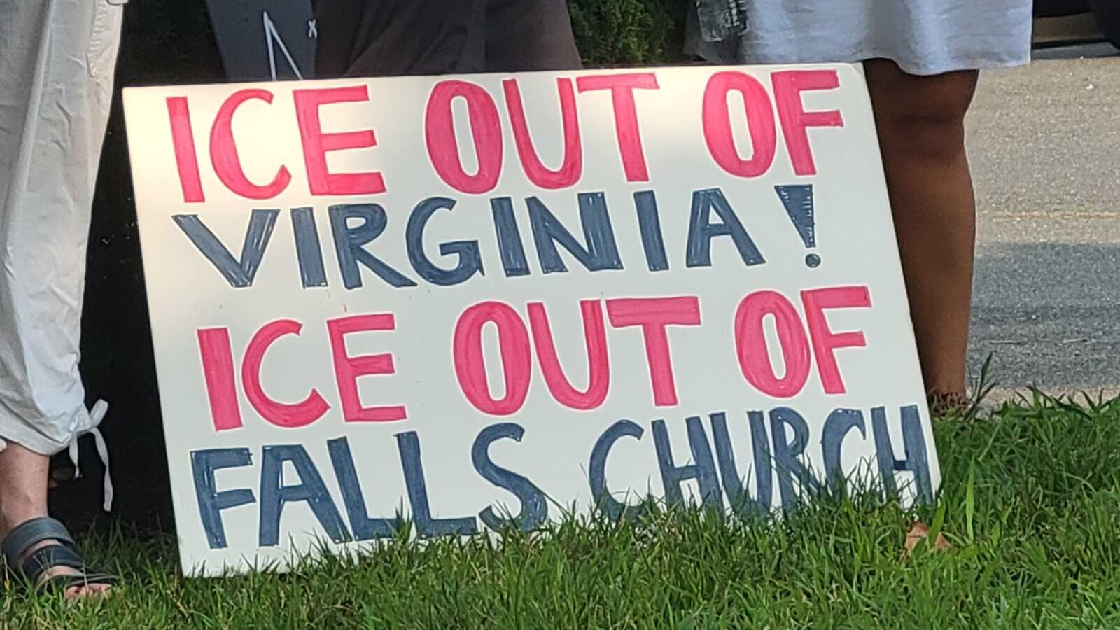 Sign reading 'ICE OUT OF VIRGINIA! ICE OUT OF FALLS CHURCH' held near a grassy curb by protesters.
