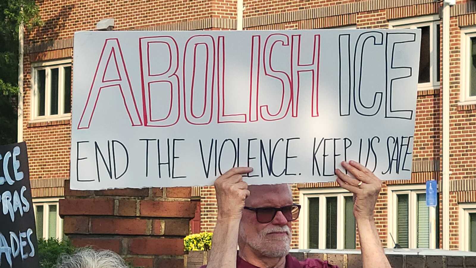 Man in sunglasses holds a protest sign reading 'ABOLISH ICE' with the subtitle 'END THE VIOLENCE KEEP US SAFE' in front of a brick building.