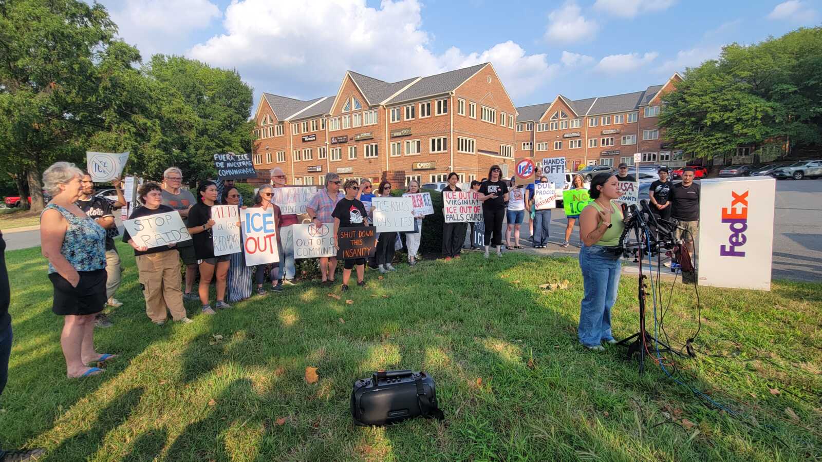 A line of diverse protesters holding hand-made signs about ICE and immigration on a grassy area in front of brick buildings, under a clear sky.