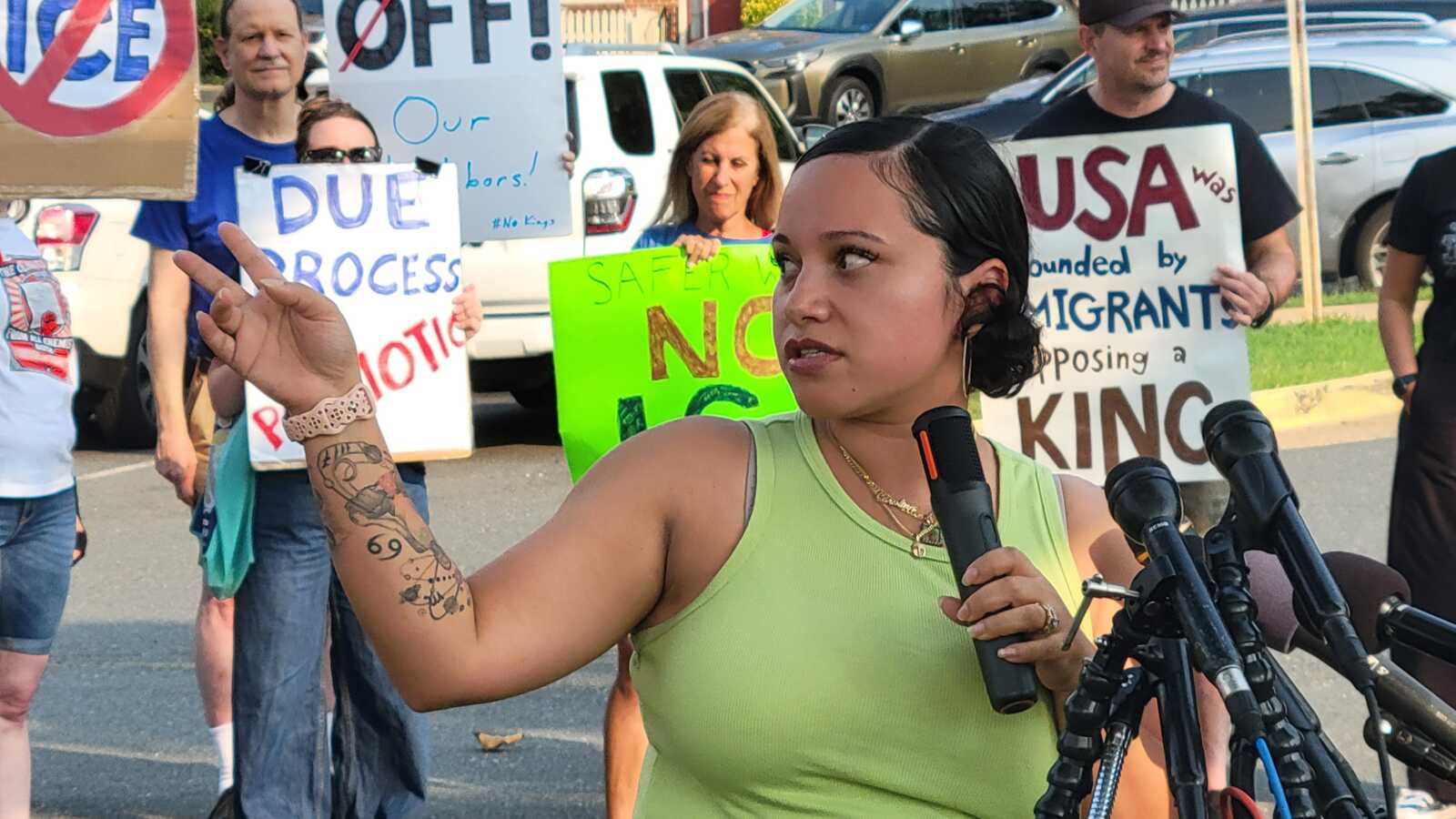 Woman in a lime-green tank top speaks into a microphone at a protest, with signs behind her, including 'USA was founded by immigrants.'