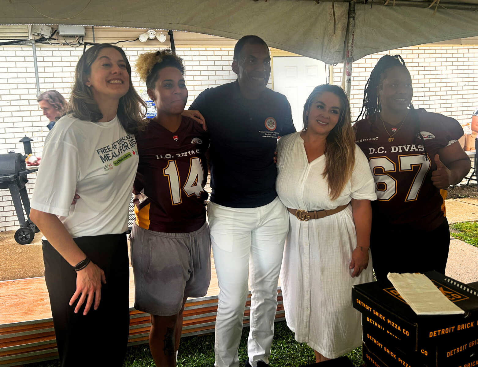Group of adults under a canopy posing for a photo at an outdoor event; two women wear D.C. Divas football jerseys (numbers 14 and 67).