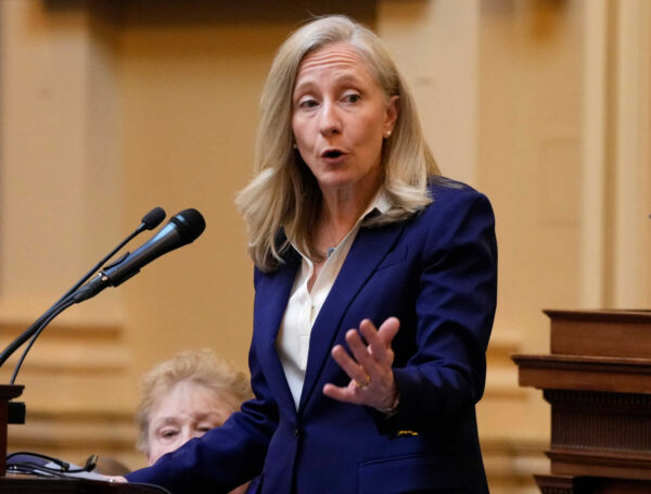 Woman in a navy blazer speaks at a podium with microphones in a formal setting.