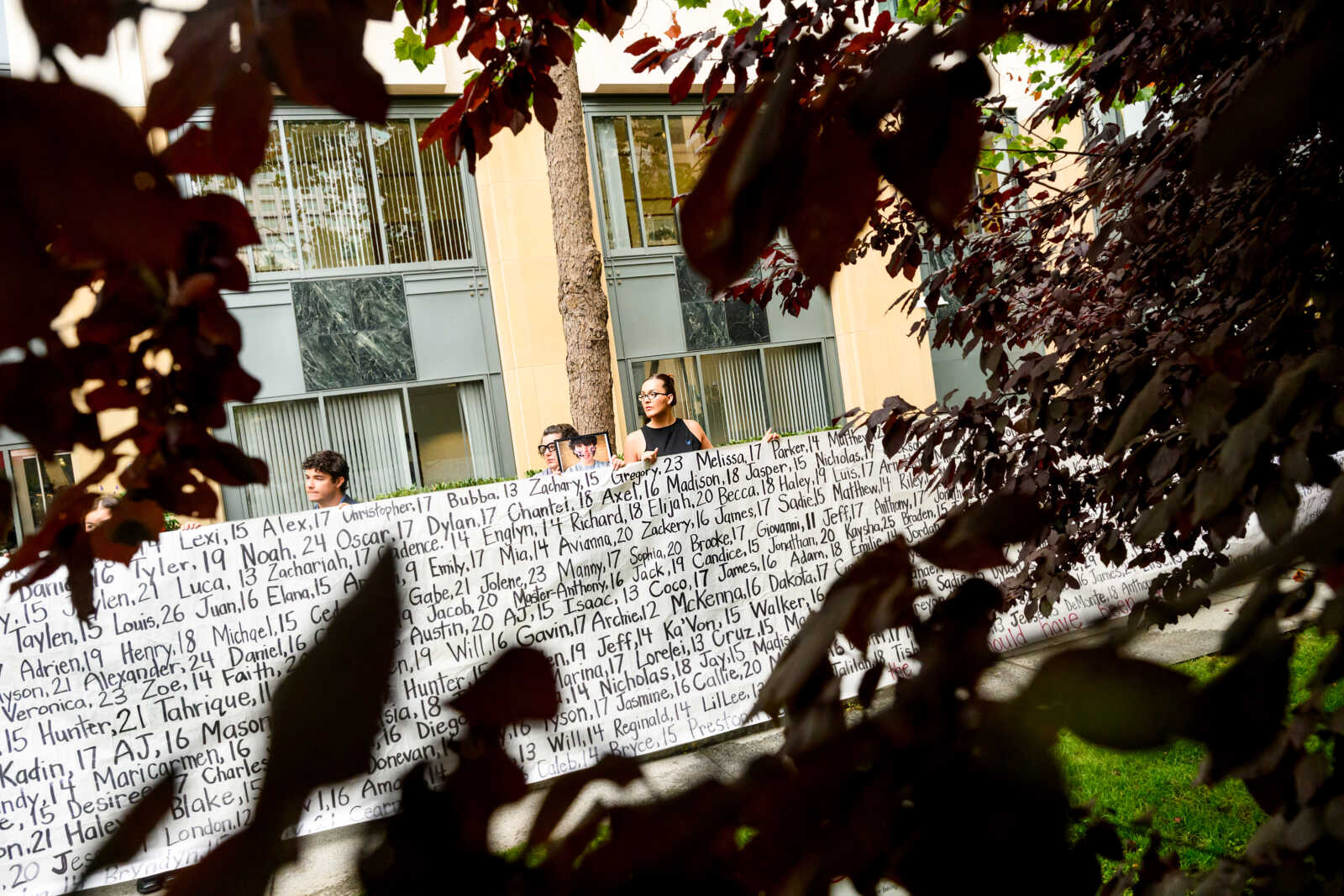 Lennon Torres of Heat Initiative holds a banner with names of young people who advocates say died as a result of social media outside the Ronald V. Dellums Federal Building and U.S. Courthouse on Tuesday, Aug. 18, 2026, in Oakland, Calif. (AP Photo/Noah Berger) | FFXnow Group of people standing behind a long wall covered with handwritten names, framed by foreground tree branches in a courtyard setting.