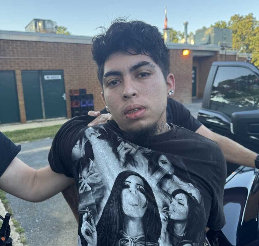 Young man with short black hair and earrings wearing a monochrome graphic t-shirt, outdoors beside a black SUV.