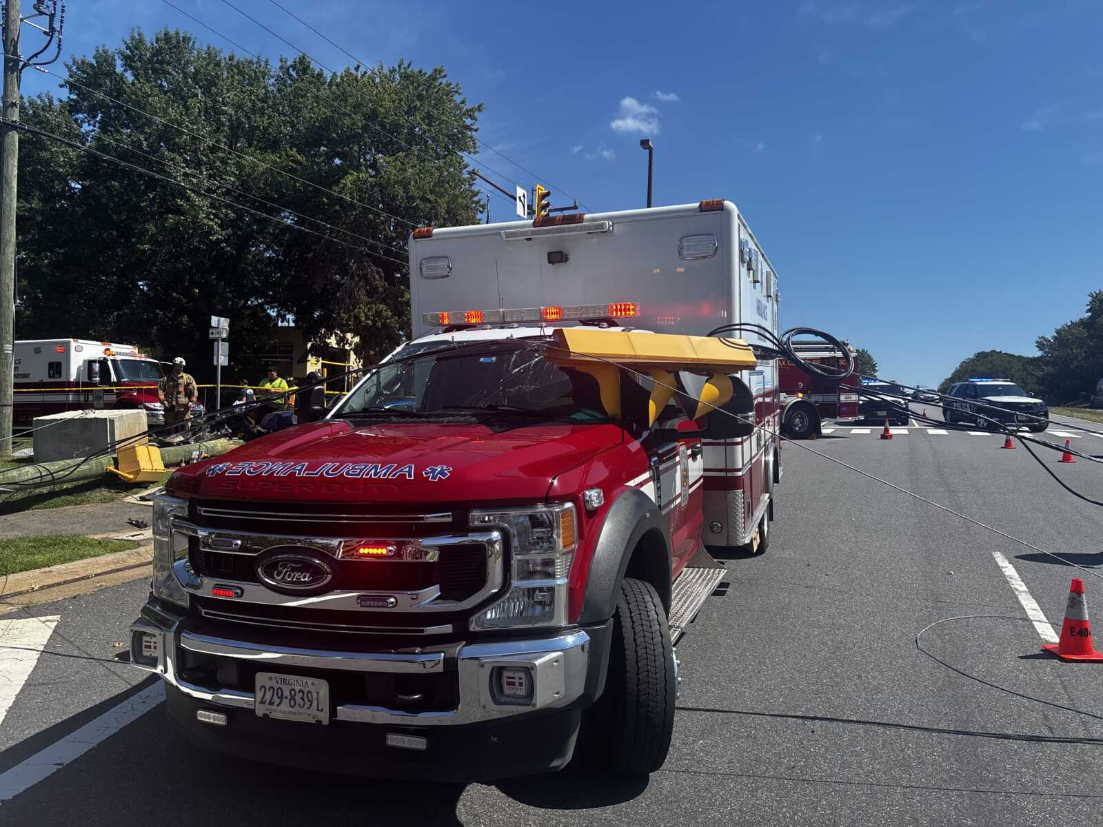 Emergency scene: a red ambulance in the foreground with responders and a crashed area on the road, police and other vehicles in the background.