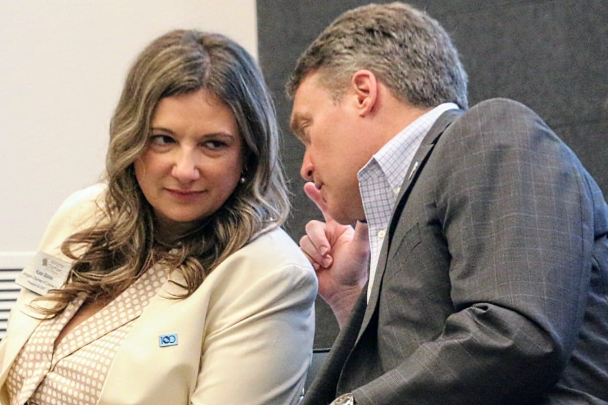 Two professionals in a meeting leaning in to discuss, the woman wearing a light blazer and the man in a gray suit.