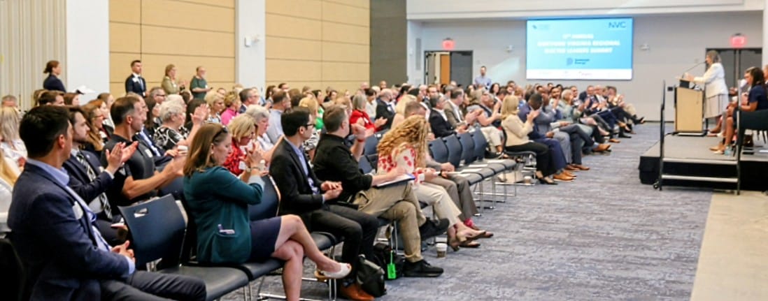 Audience at a conference applauds while a speaker presents on a stage with a podium and a screen in the background.