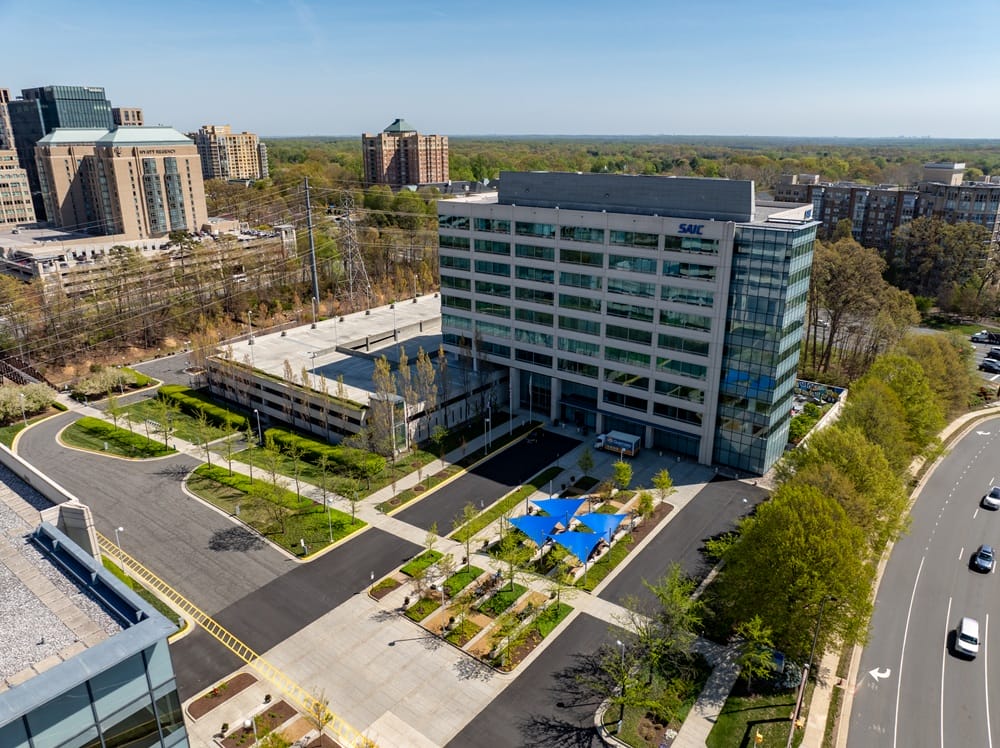 The office building at 12010 Sunset Hills Road in Reston Town Center (courtesy BXP) | FFXnow Aerial view of a modern office campus with a tall glass-and-concrete SAIC building, surrounding parking, and trees.