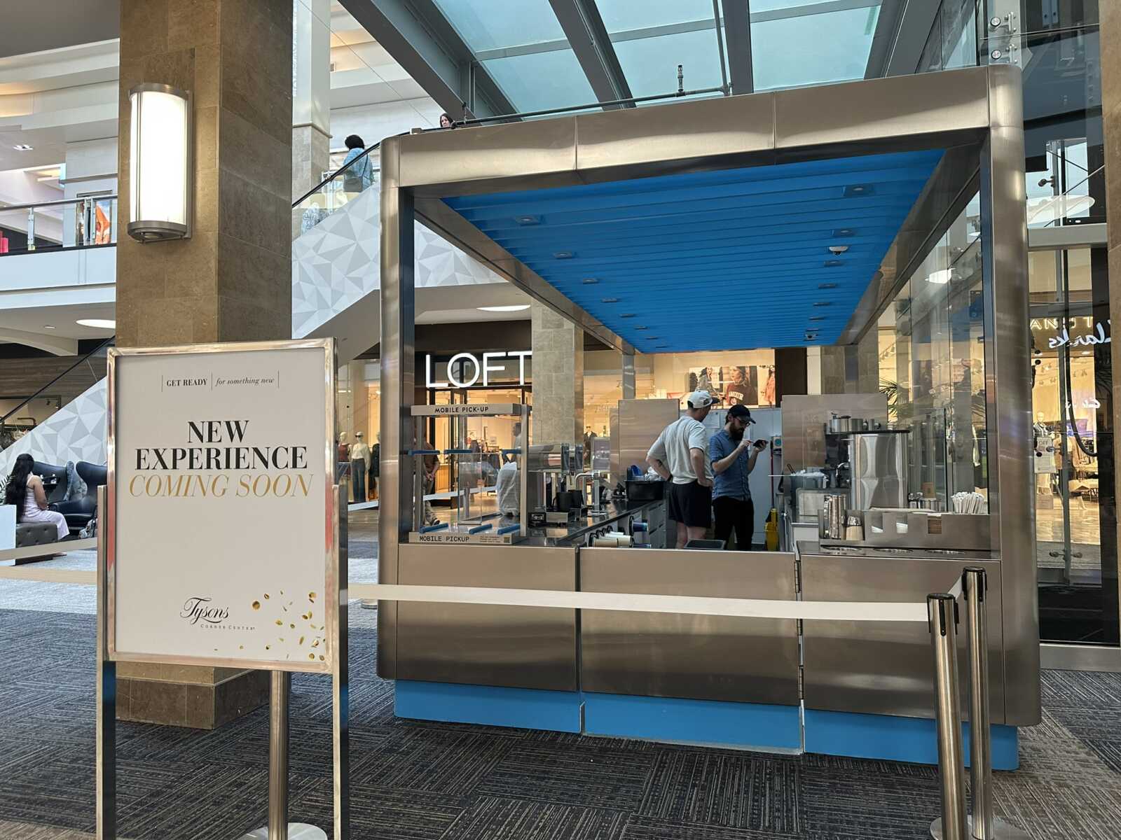 Mall kiosk with two staff at a stainless steel counter under a bright blue ceiling, LOFT store in back, and a sign reading 'New Experience Coming Soon' nearby