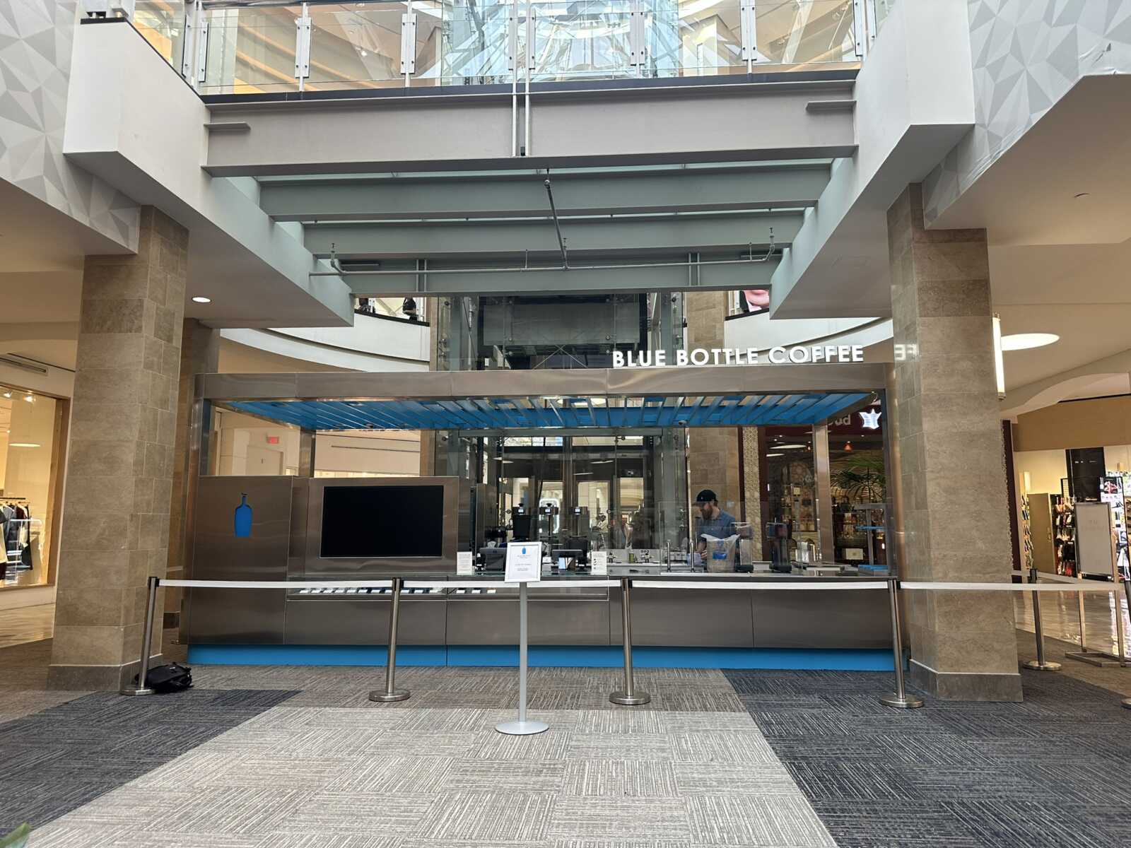 Blue Bottle Coffee kiosk in a mall atrium with a glass railing above and a service counter behind a barrier.