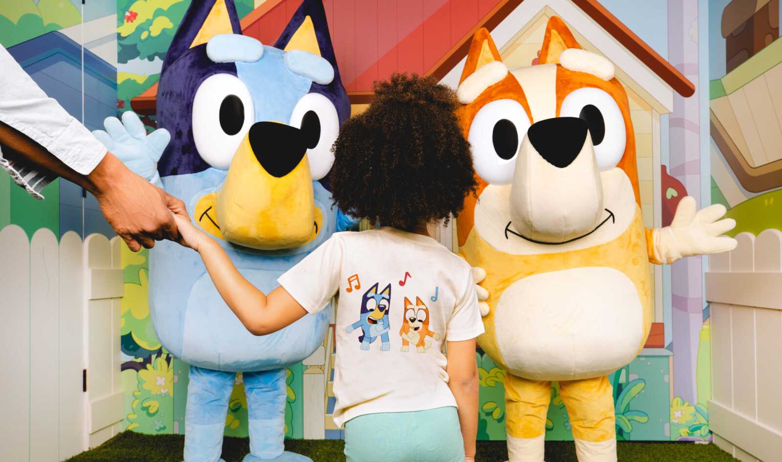 A child in a white t-shirt with cartoon dogs stands between two large colorful mascot characters, blue on the left and orange on the right, as a hand reaches in from the left to join the hug.