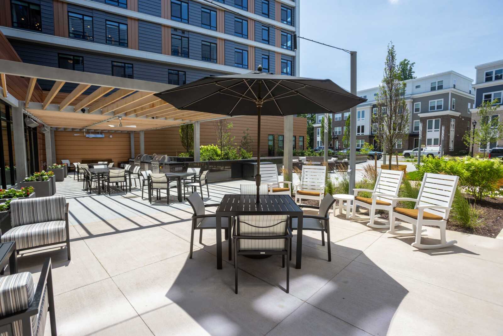 Outdoor seating area of a modern apartment complex with tables, chairs, and a large black umbrella under a pergola.