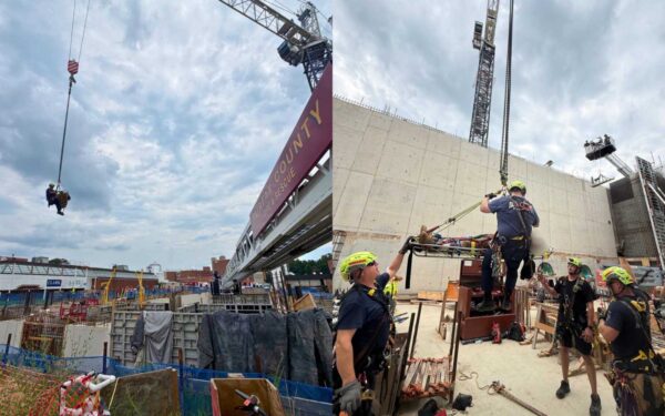 Rescue team hoists a person in a basket with a crane over a construction site, workers nearby in safety gear.