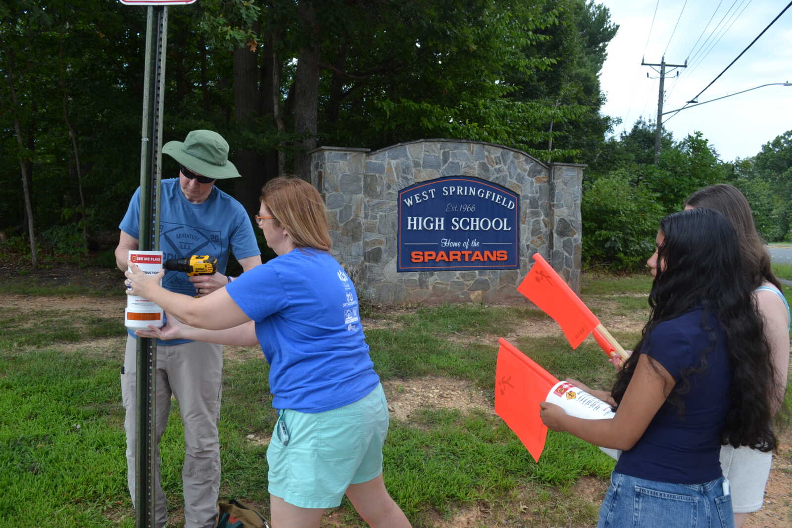 People installing a bucket near a stone sign that reads West Springfield High School, Home of the Spartans, with orange flags nearby in a grassy area.