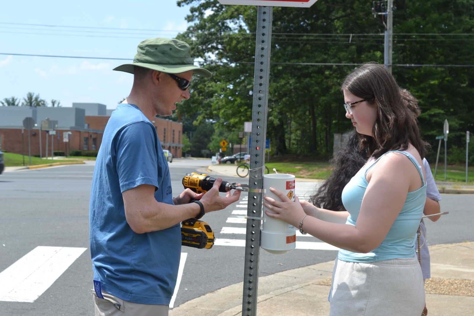 Two people work together on a street corner: a man with a drill attaches a device to a metal sign post while a young woman holds a white cylindrical unit at the base, near a crosswalk.