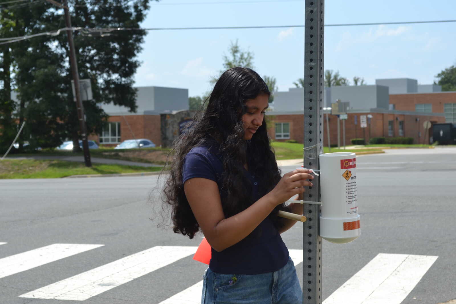 Young woman with long dark hair stands at a crosswalk beside a metal street sign post.