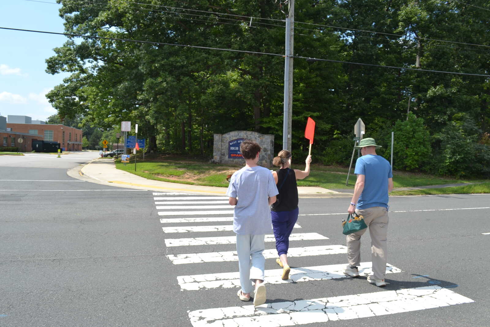 Three pedestrians cross toward a sidewalk with trees and a stone sign in the background on a sunny day.