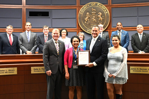 Officials pose in a government chamber as a man presents a framed certificate to a woman in front of a large County of Fairfax seal and podiums.