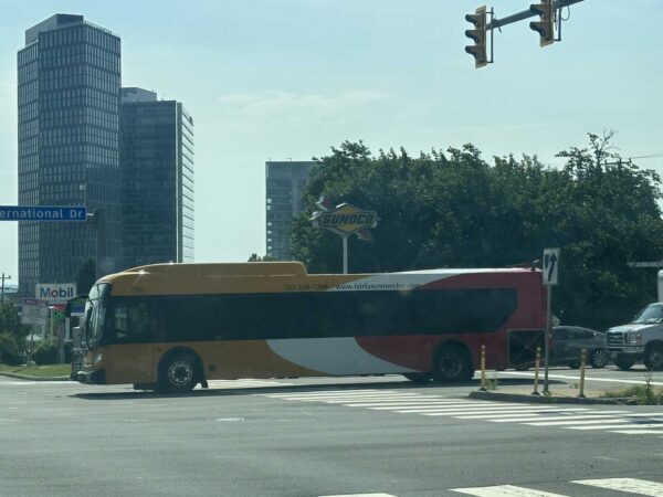 Yellow and red tour bus crossing an urban intersection under a traffic light with tall office buildings in the background and a Sunoco sign nearby.