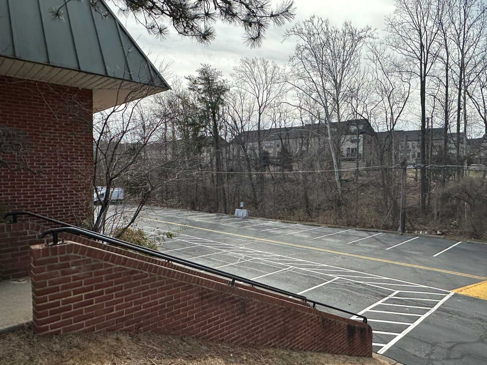 Brick building corner with metal roof overhang, brick railing, and a mostly empty parking lot beyond with numerous painted lines and leafless trees in the background on a cloudy day.