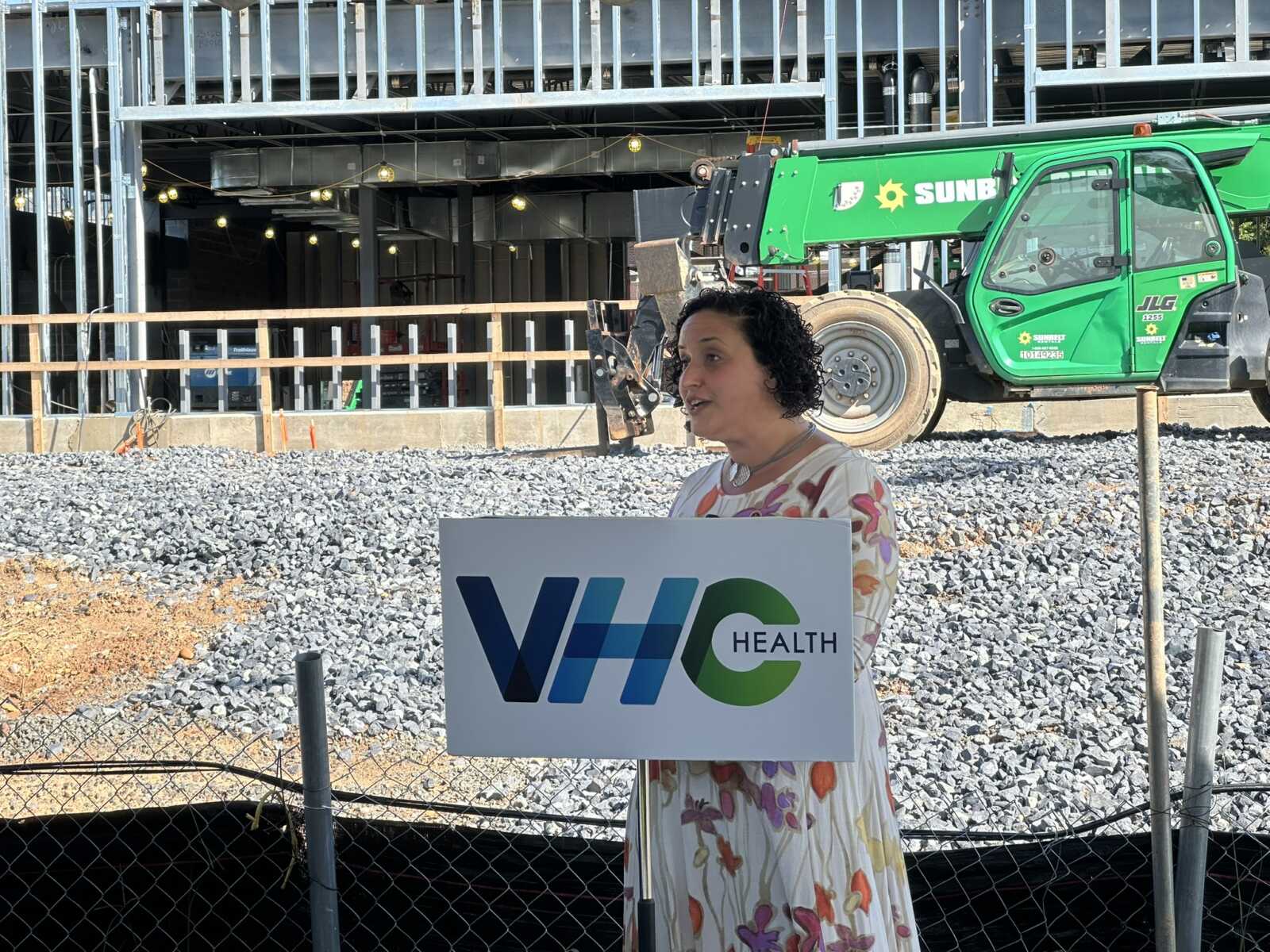 Woman speaking at a VH Health podium at a construction site, with a green crane in the background.