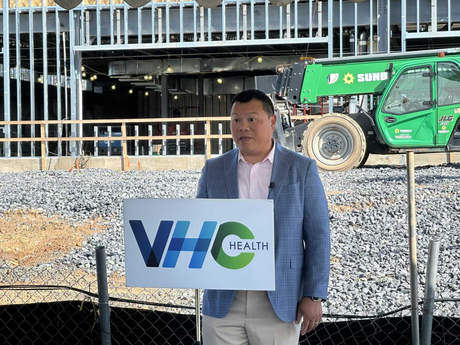 Man in a blue blazer speaks at a construction site, standing behind a WHC Health podium with a green crane in the background.
