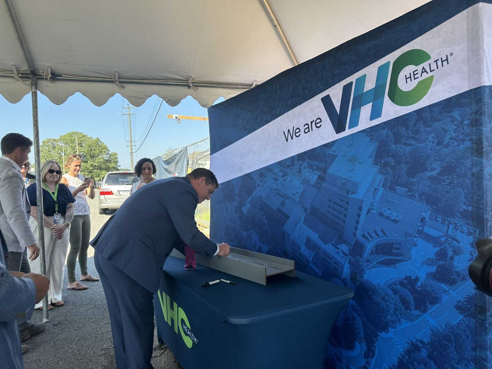 Man in a suit signs a document at a VHC Health booth under a white tent while several people watch nearby, next to a large 'We are VHC Health' banner.