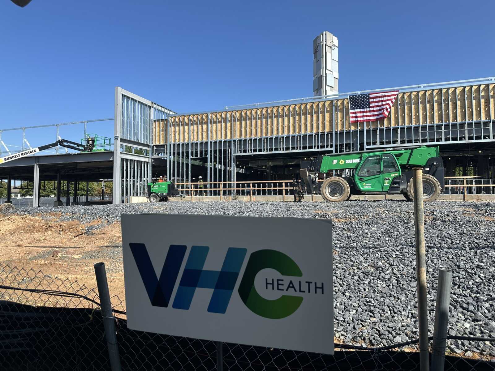 Construction site for a health facility with steel framing, a green lifting machine, and an American flag on the building's exterior.
