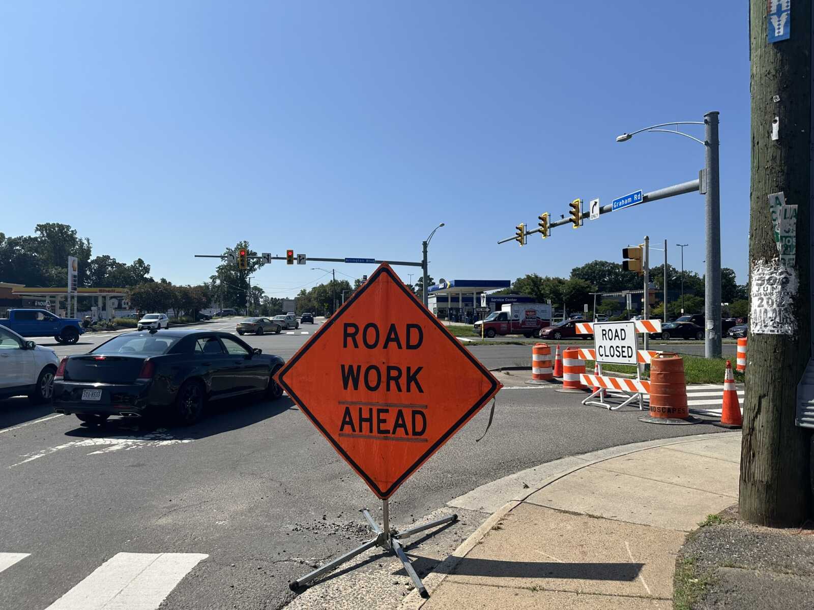 Orange diamond 'ROAD WORK AHEAD' sign in the foreground at a busy intersection with traffic and construction barrels nearby, indicating road work ahead.