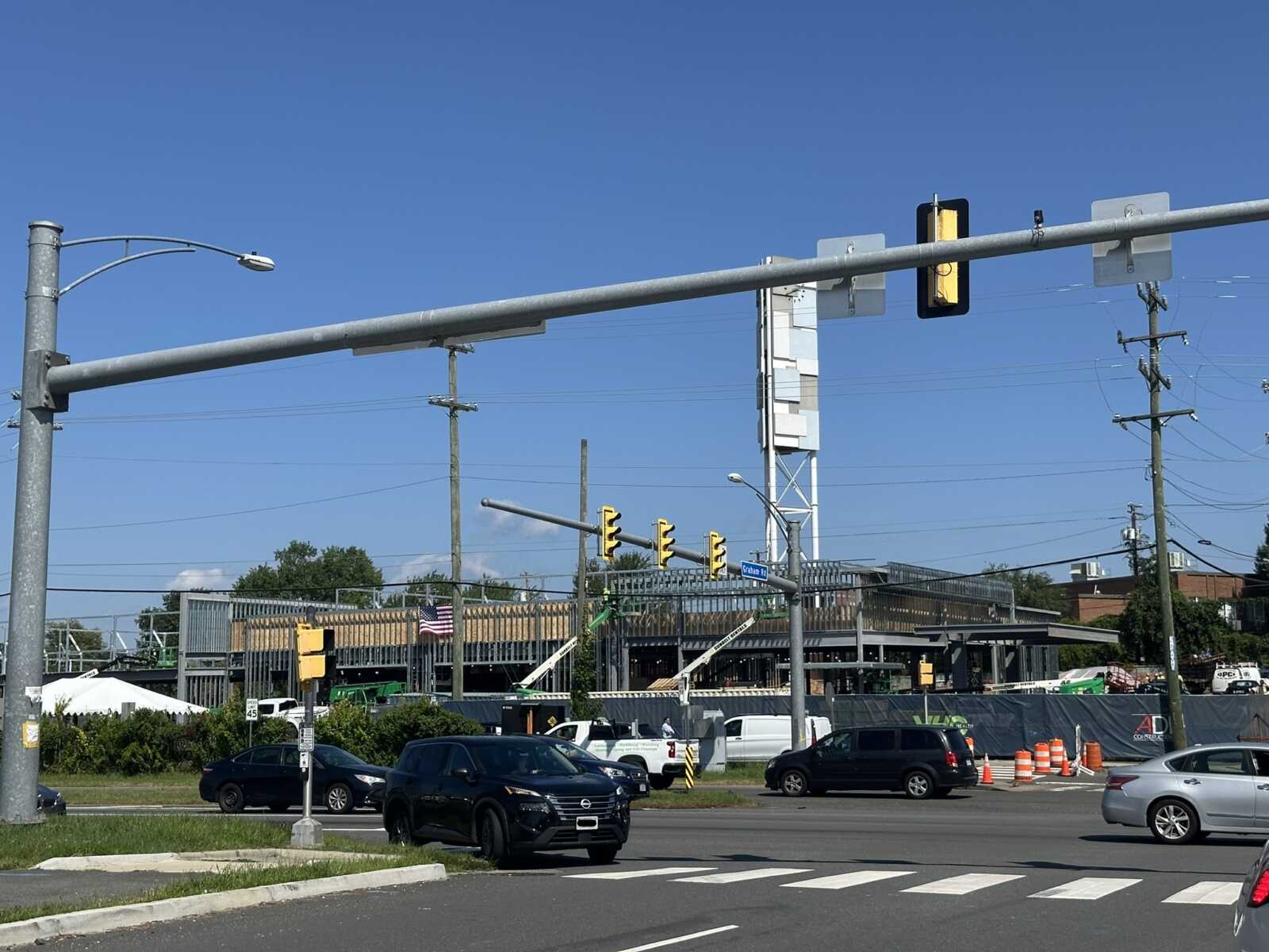 VHC Health and Intuitive Health's combined emergency and urgent care center, now under construction, in West Falls Church (staff photo by Angela Woolsey) | FFXnow Busy urban intersection under construction with barriers, parked cars, and overhead traffic signals against a clear blue sky.
