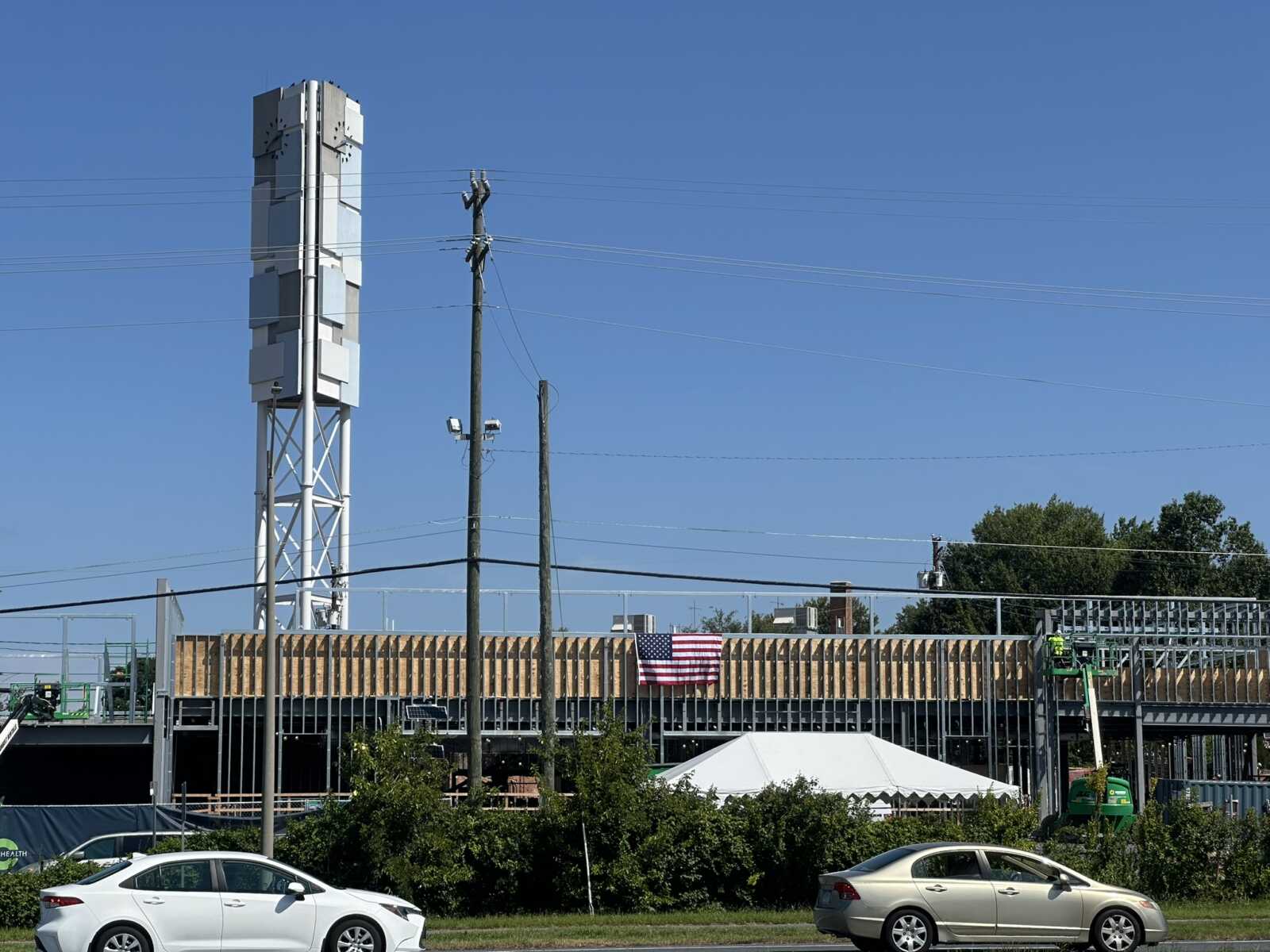 Construction site along a road with a tall white telecom tower, power lines, and wooden framing under clear blue skies with an American flag nearby.