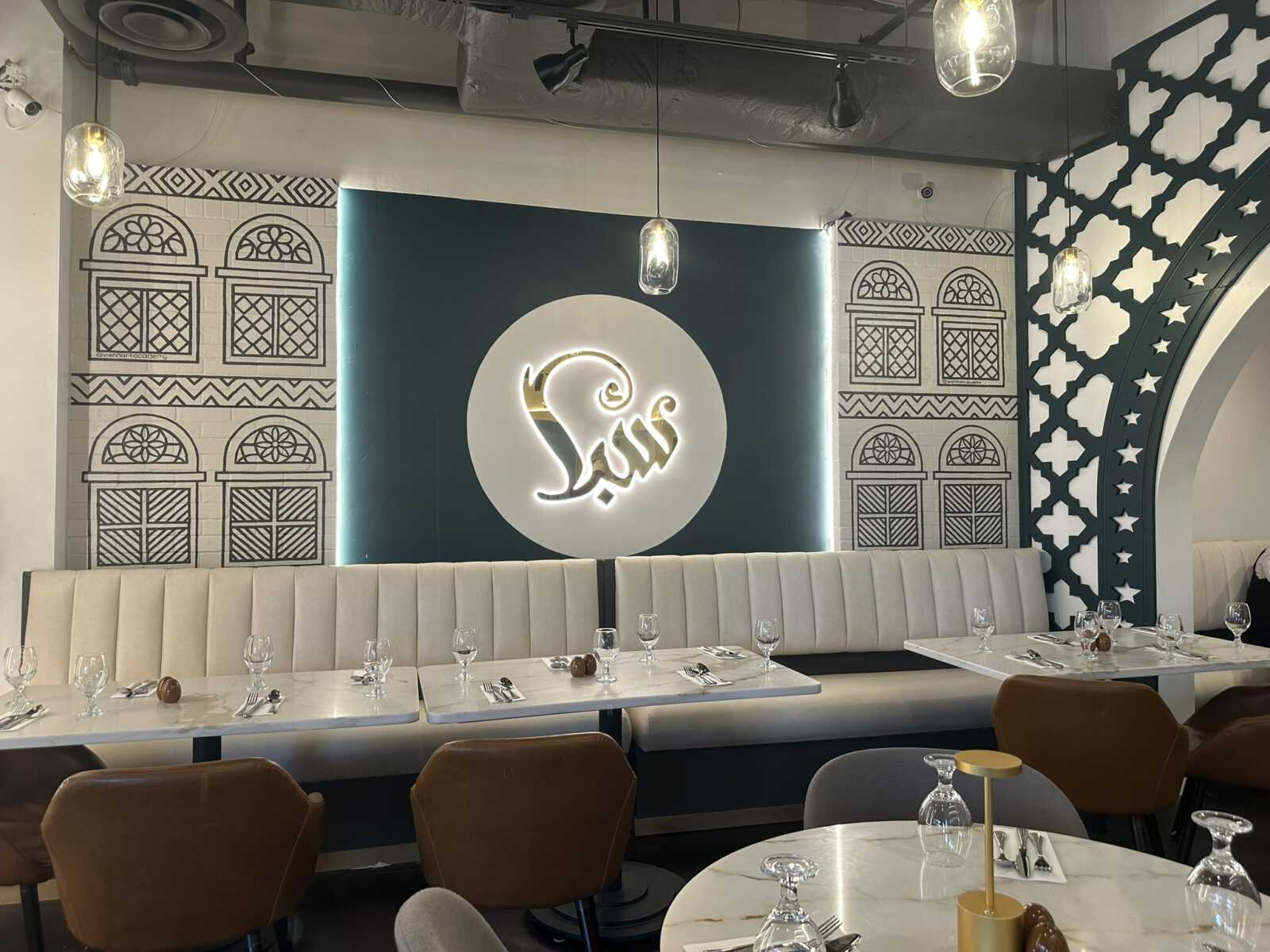 Dining area with white upholstered banquette, round marble tables, and glassware; teal wall features a circular illuminated logo with Arabic script at center.