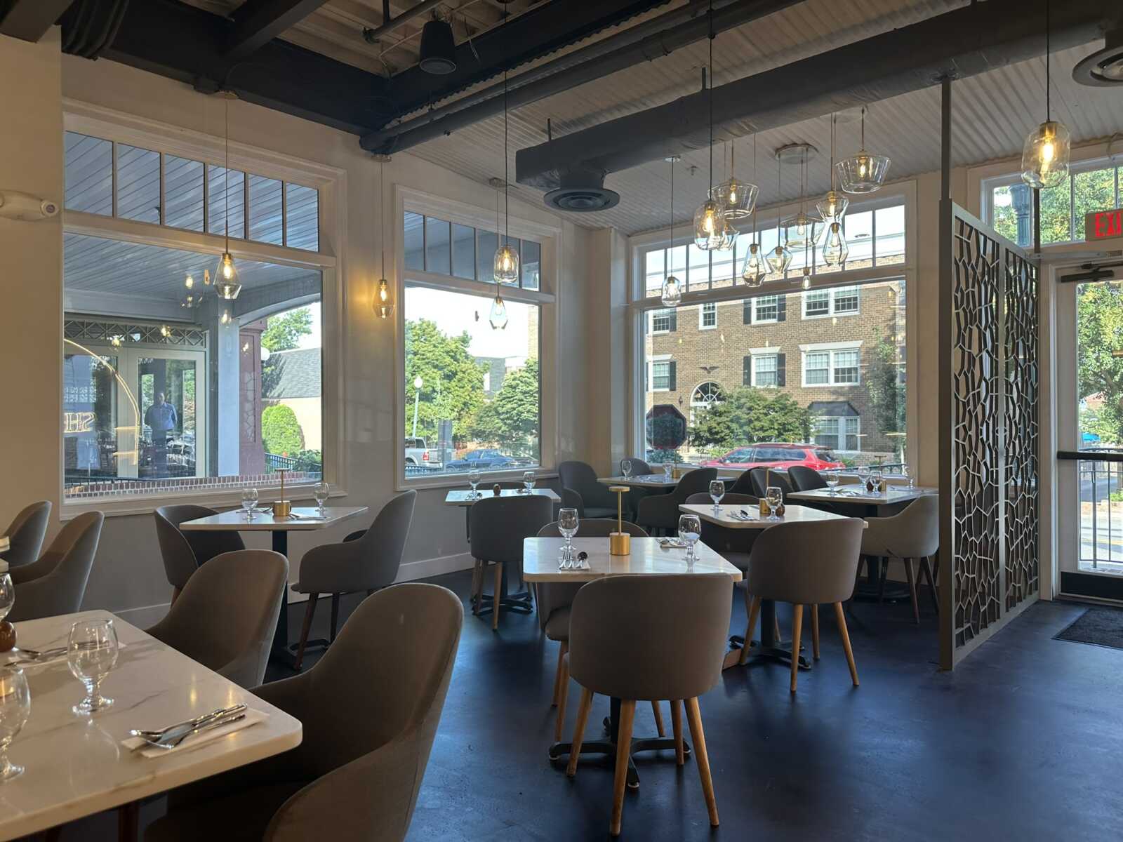 Modern restaurant dining area with white tables, beige chairs, large windows, and pendant lights overhead at dusk.