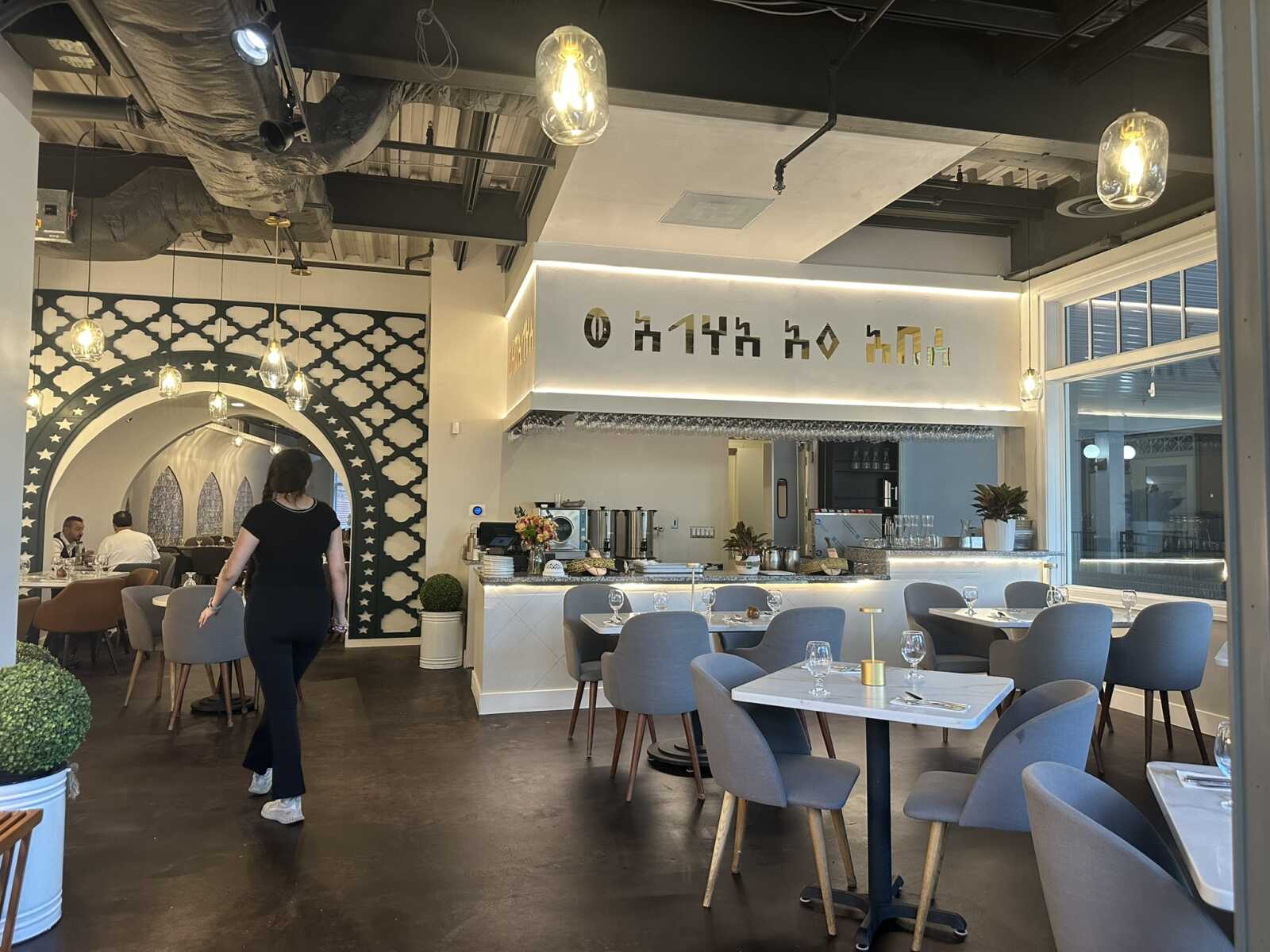 Modern restaurant interior with blue-gray chairs, white tables, and a bar area; decorative arched wall with geometric patterns on the left.