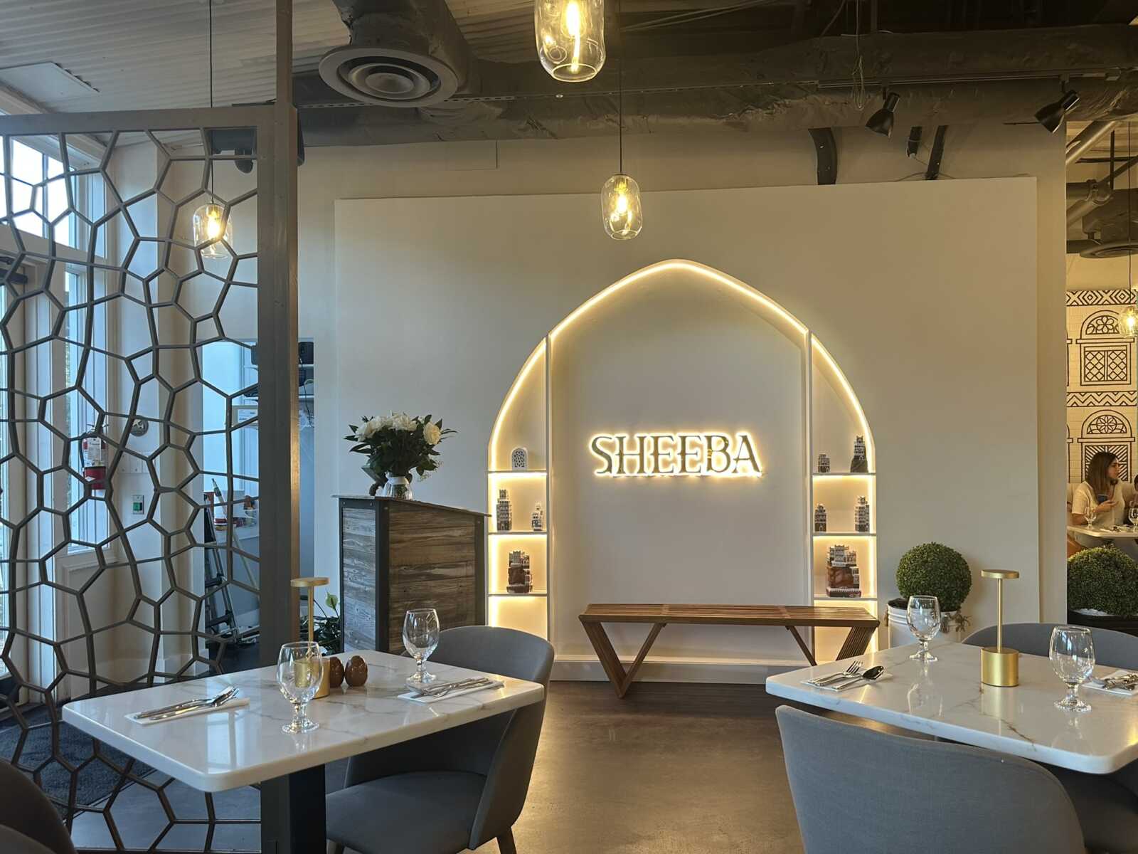 Modern restaurant interior with a glowing arched display reading 'SHEEBA' and lit shelves along the wall, marble tables and pendant lights.