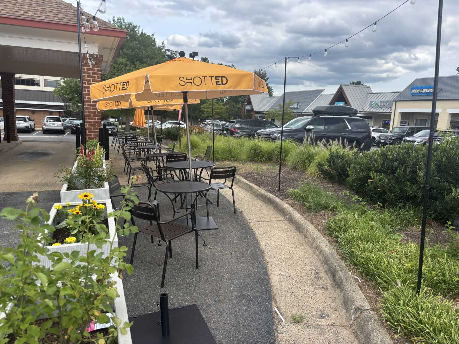 Outdoor cafe seating with orange umbrellas and black metal tables along a sidewalk, planters, and string lights overhead in a shopping center.