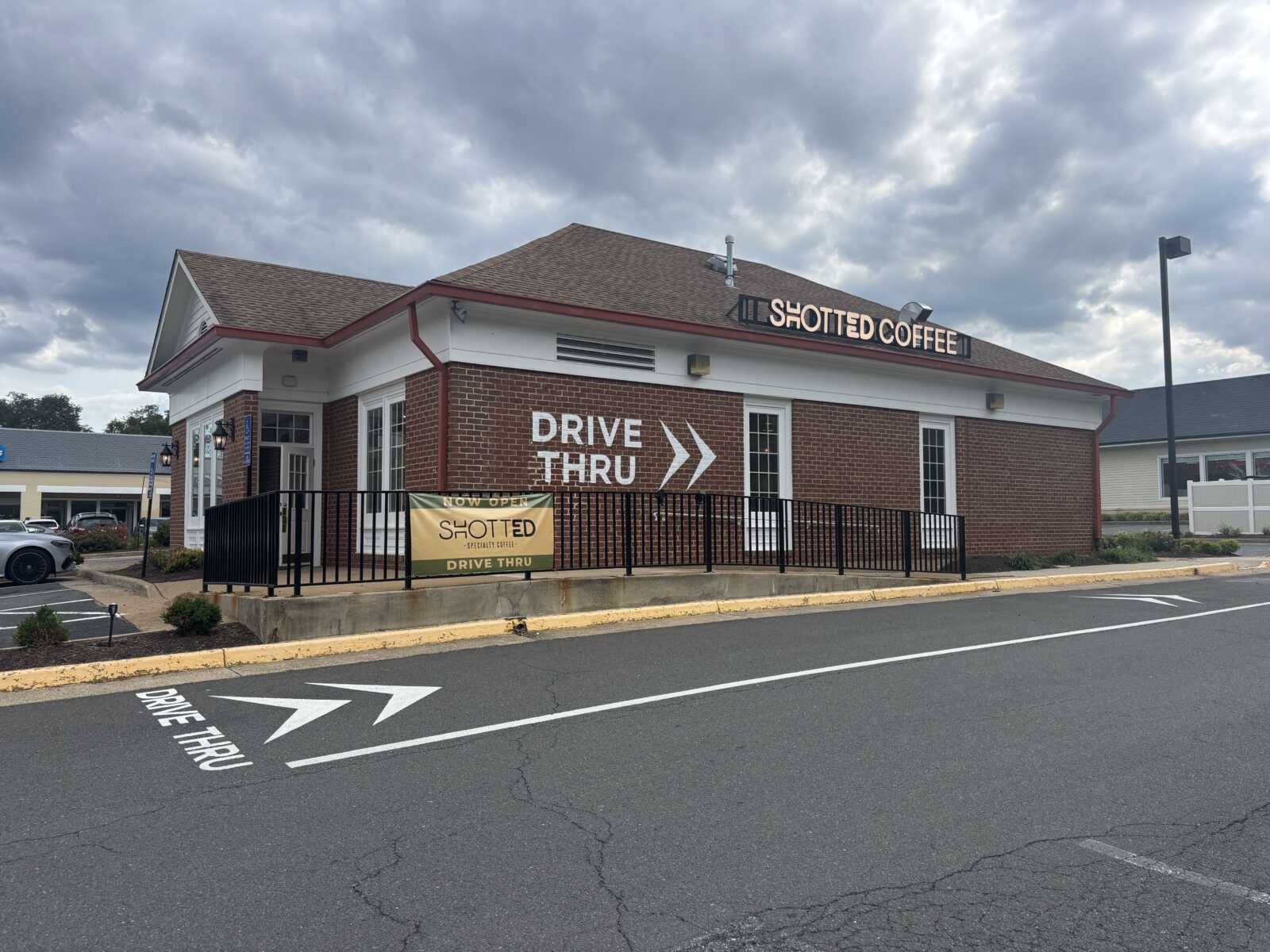Exterior of Shotted Coffee storefront: brick building with white trim, drive-thru arrows on the wall, and a banner along the railing announcing the opening.