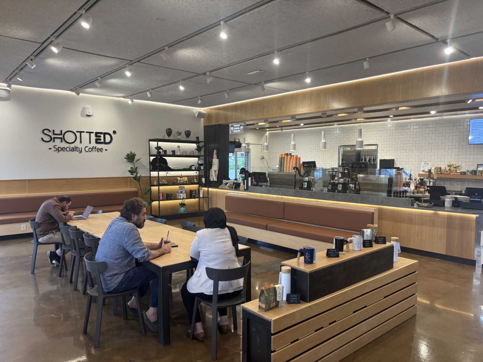 Modern coffee shop interior with a long wooden communal table and seating, customers working and chatting; bar counter in the background with coffee machines and menu boards.