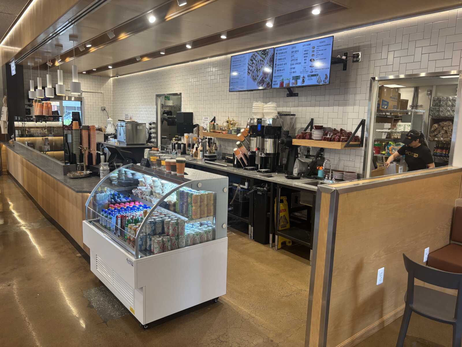 Coffee shop counter with espresso machines, stacked cups, and digital menu boards above the bar area.