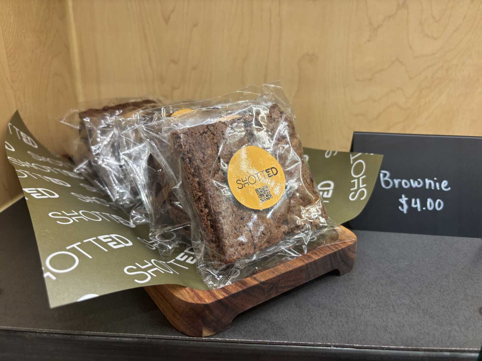 Brownie wrapped in plastic with an orange circular label on a wooden display, background shows a sign reading 'Brownie $4.00'.