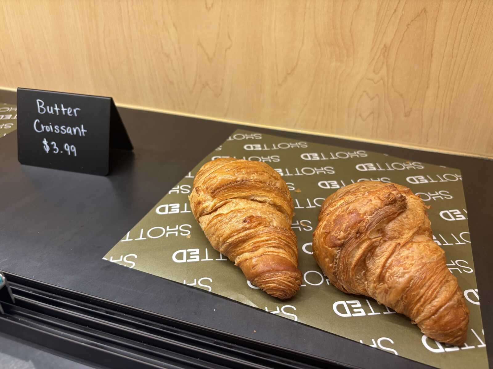 Two buttery croissants on a branded display mat with a price sign reading ‘Butter Croissant $3.99’ in the background.