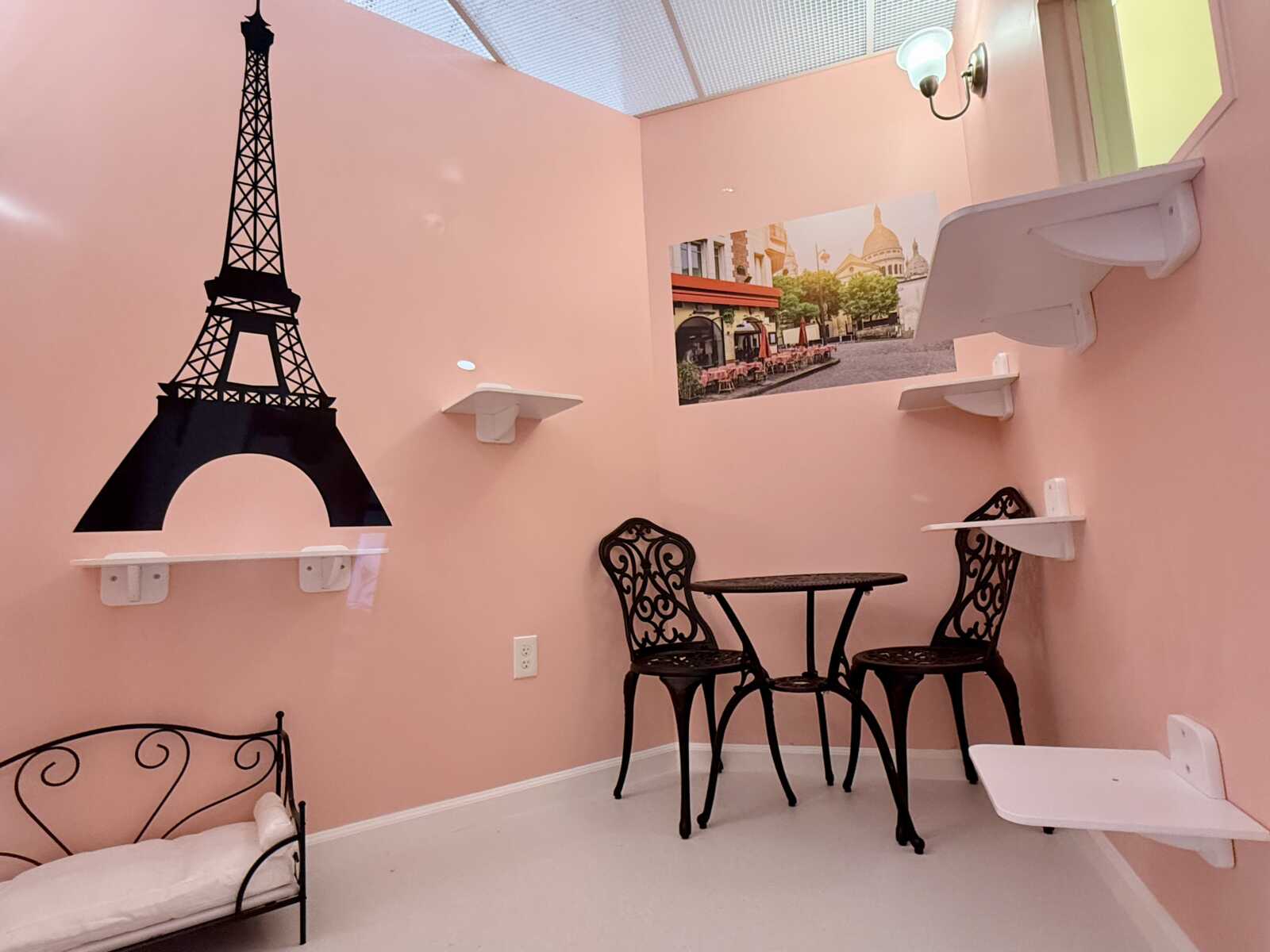 Pink room with a black Eiffel Tower wall decal, a small round table and two ornate metal chairs, and white wall shelves and mural.