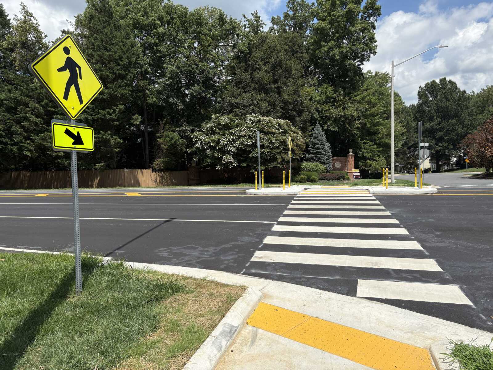 Crosswalk with wide white stripes across a road, two bright yellow pedestrian warning signs on the left, and trees in the background.