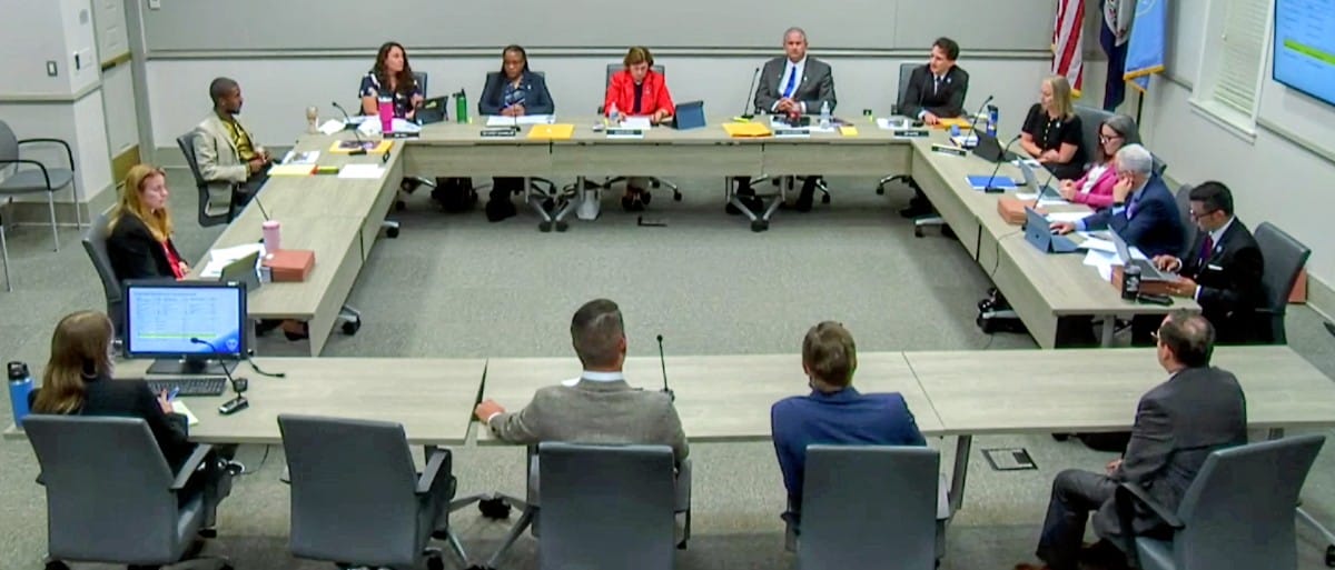 Formal board or council meeting around a U‑shaped table with multiple participants, laptops, papers, and microphones in a conference room.