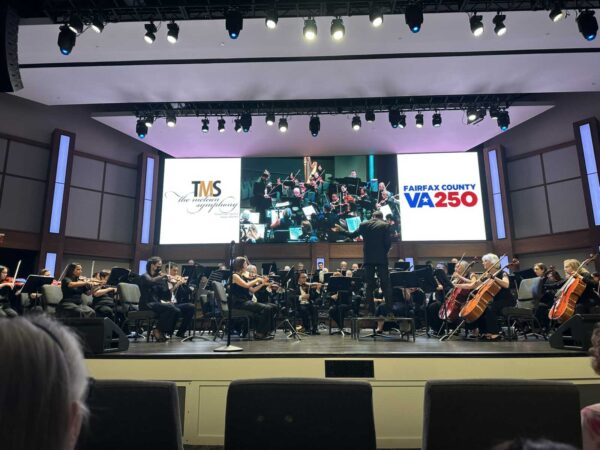 Orchestra on stage with a conductor, large screens behind showing logos for TMS and Fairfax County VA-250, in a concert hall.