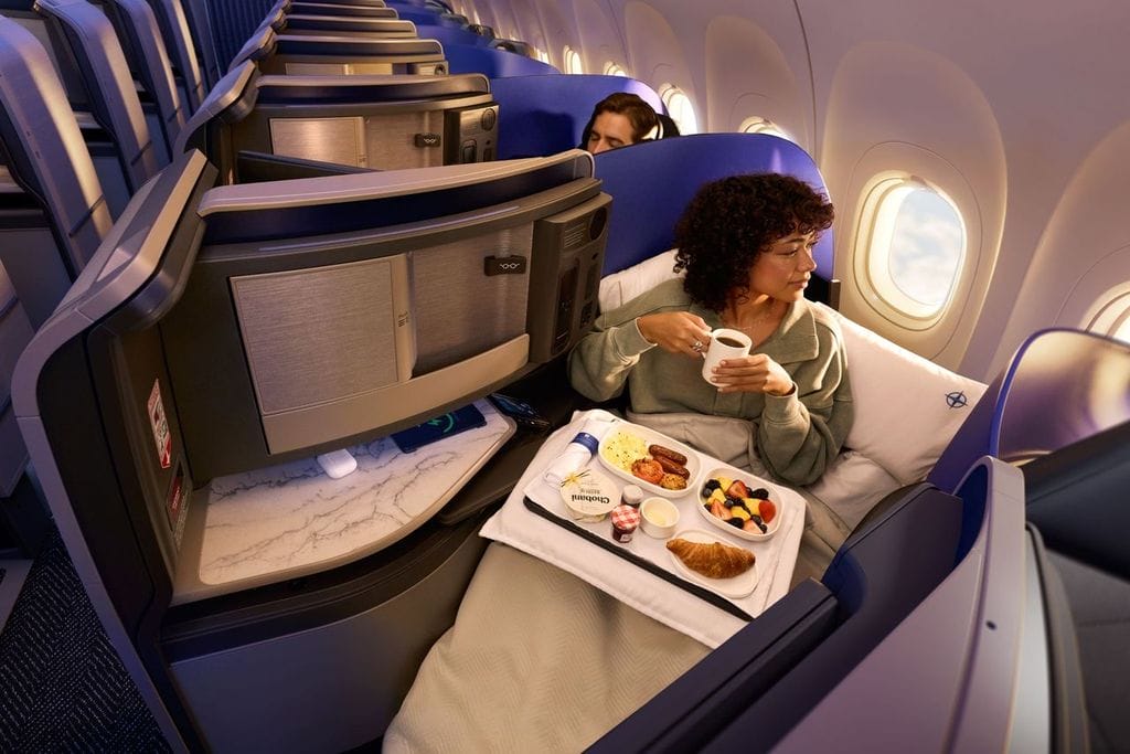 Woman in a business-class seat sipping coffee with a tray of breakfast foods beside her at flight’s edge seating.