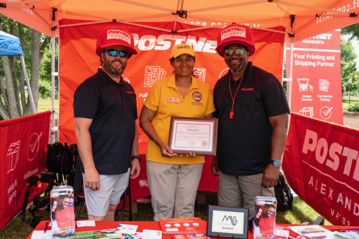 Three people pose in a PostNet booth at an outdoor event; a woman in a yellow shirt holds a framed certificate between two men wearing red hats.