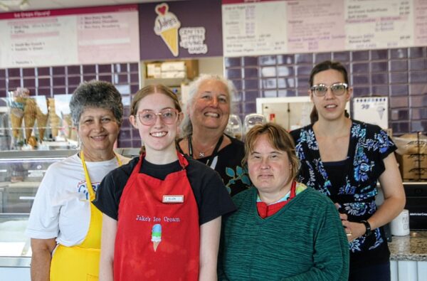 Group of five smiling staff posing behind an ice cream counter in a shop, one wearing a red apron that says 'Jake's Ice Cream'.