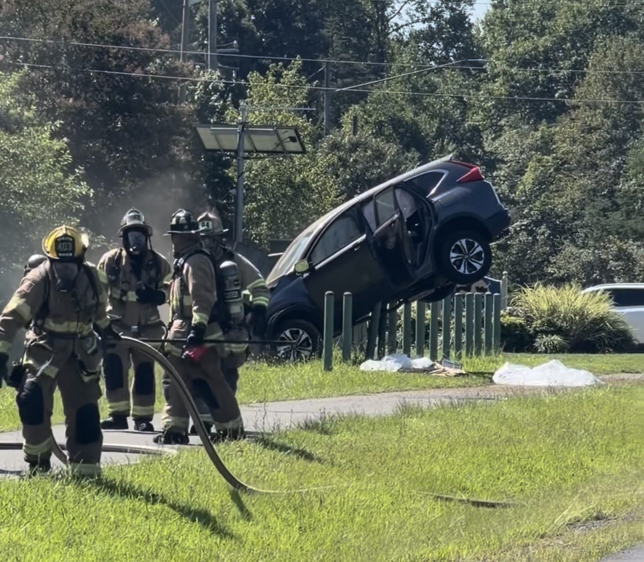 Firefighters in full turnout gear spraying a vehicle crash scene; a dark SUV is lodged on a wooden fence at the roadside, with smoke in the background.