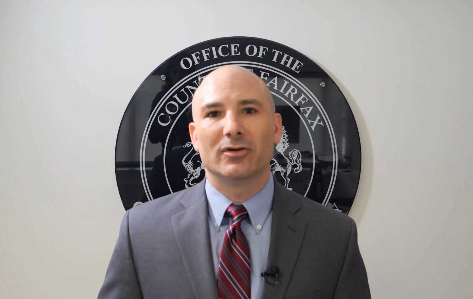 Man in a gray suit and red striped tie speaks to camera, standing in front of a large circular seal on the wall behind him (office setting).